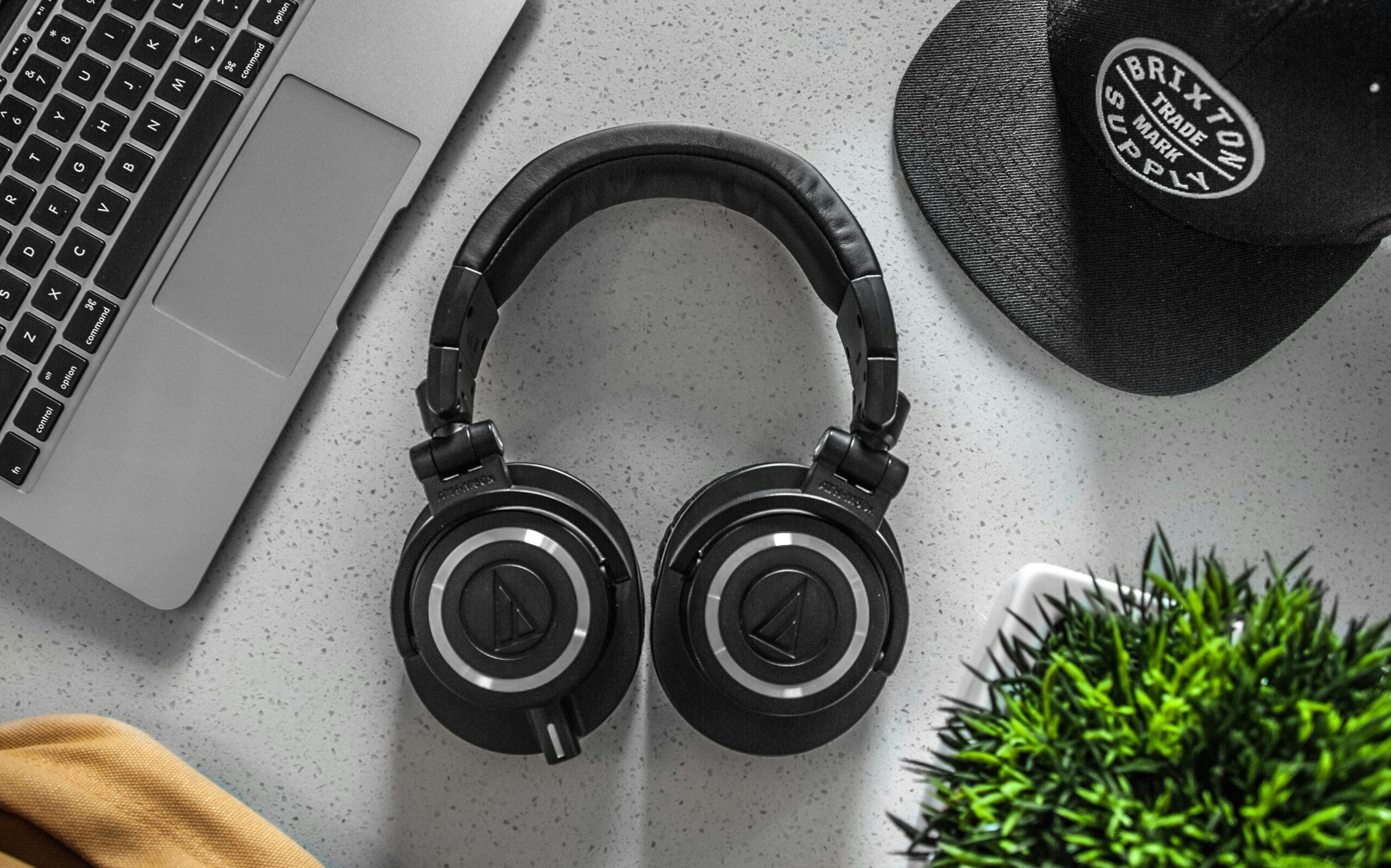 A stylish overhead shot of headphones with a laptop, cap, and decor on a desk.